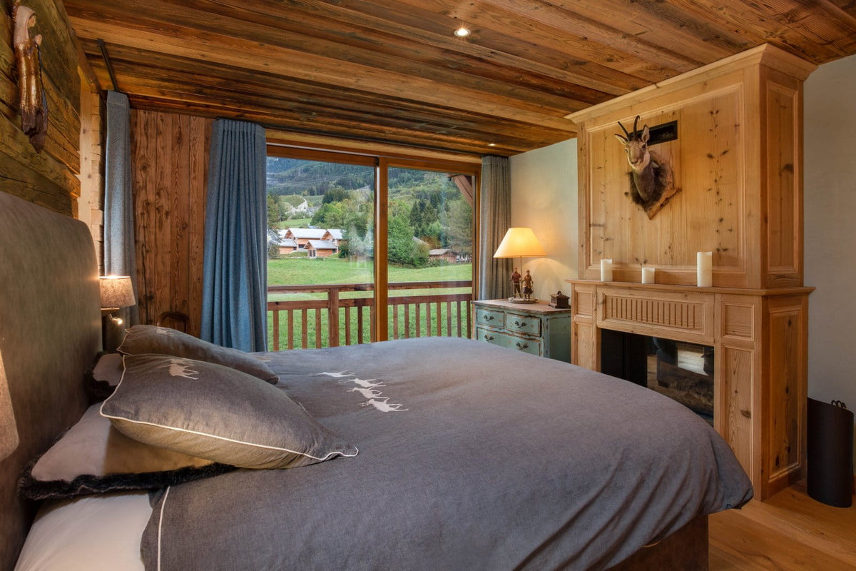 A cozy bedroom interior with a large bed dressed in gray bedding, set in a rustic wooden cabin with a fireplace, blue curtains, and a window overlooking a green outdoor landscape.