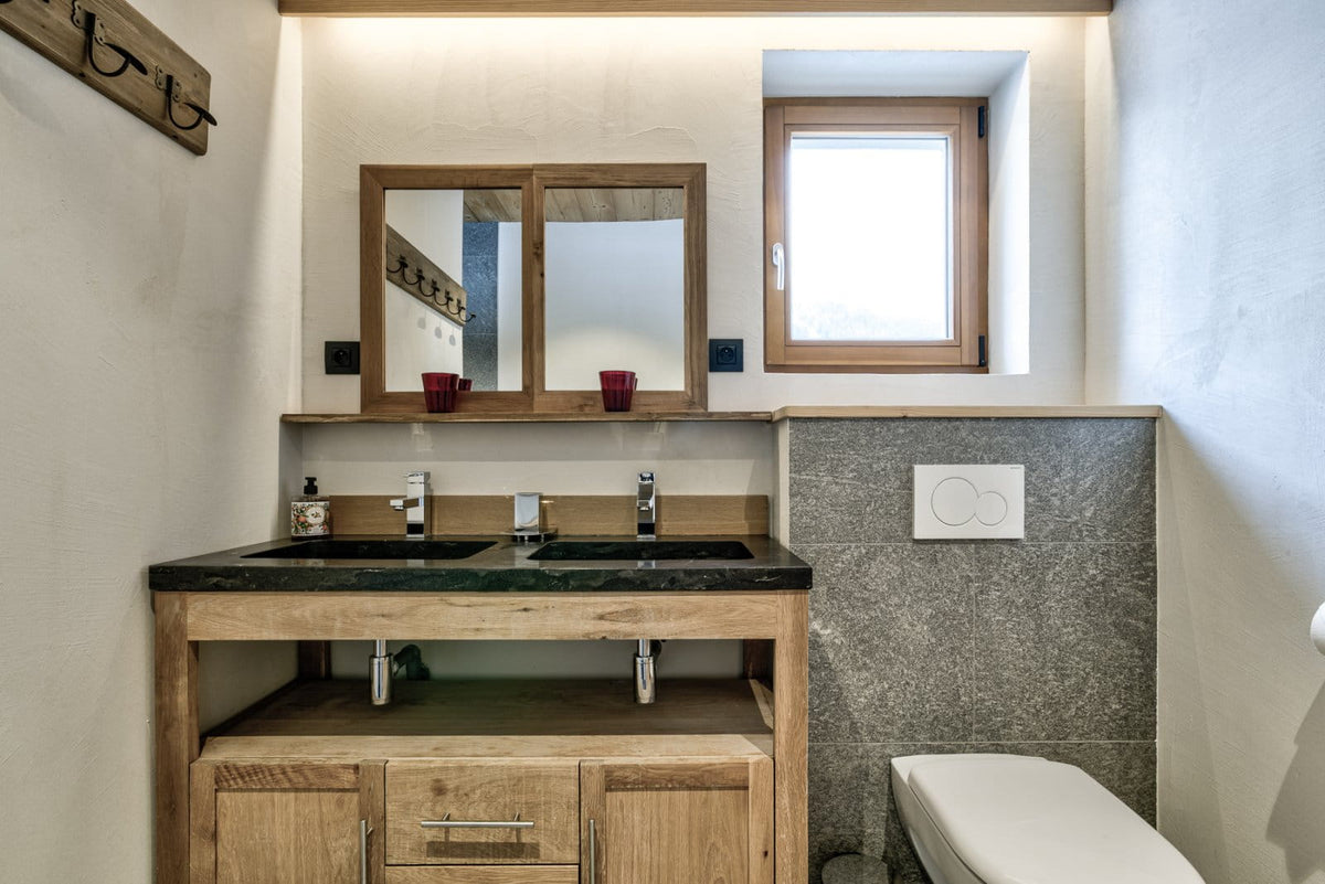 A rustic double-sink bathroom vanity made of light natural wood, featuring a dark countertop, a matching wood-framed mirror, and a textured gray stone backsplash.