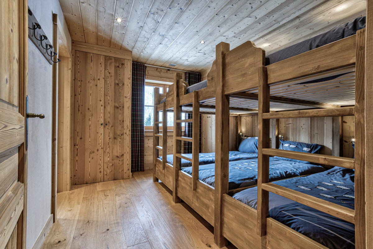 A rustic wooden bedroom with a set of three wooden bunk beds dressed in blue patterned bedding, set against light wood paneled walls and ceiling.