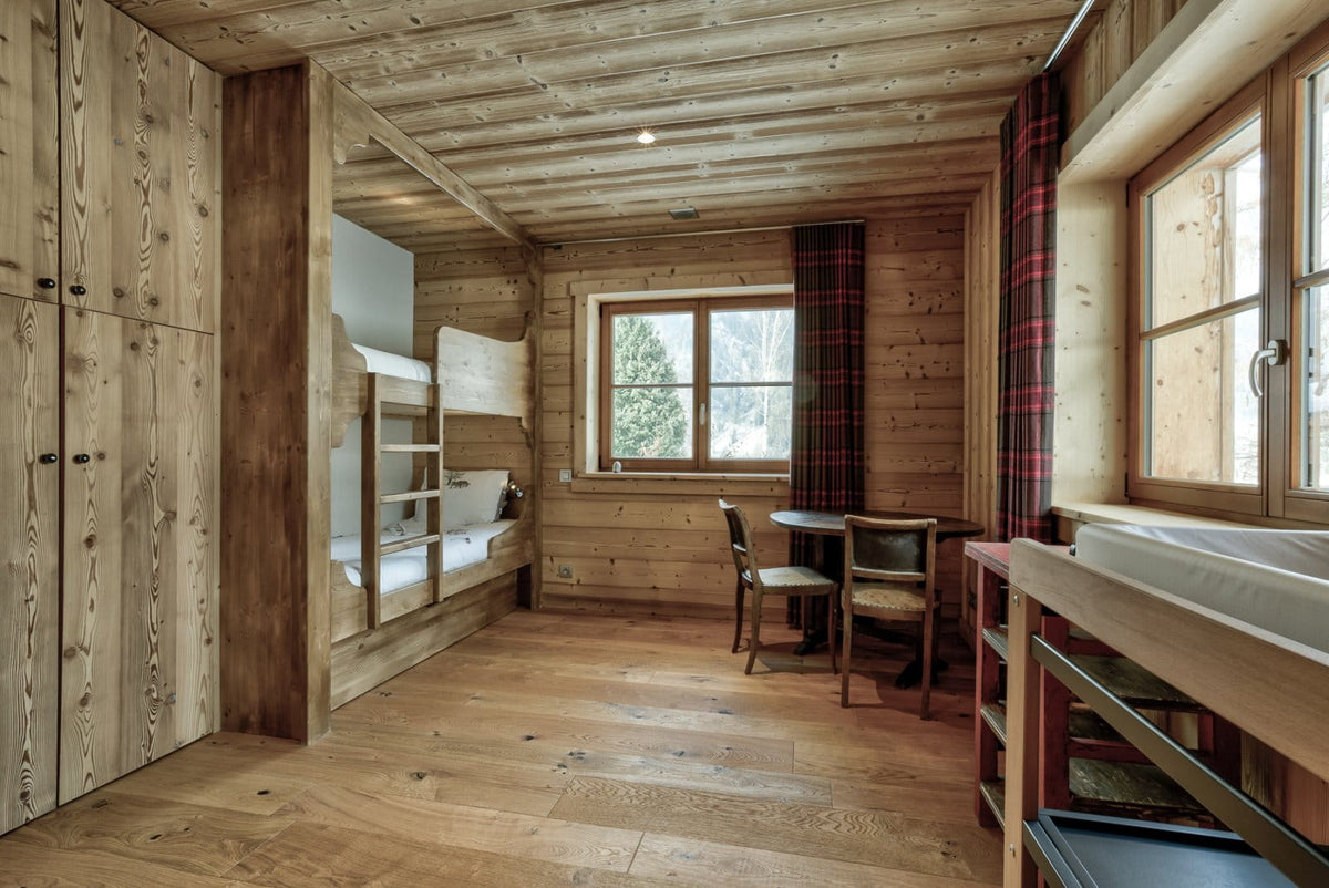 A rustic wooden chalet interior featuring a built-in wooden bunk bed, a small wooden desk with two chairs, and red-and-black plaid curtains by a window.