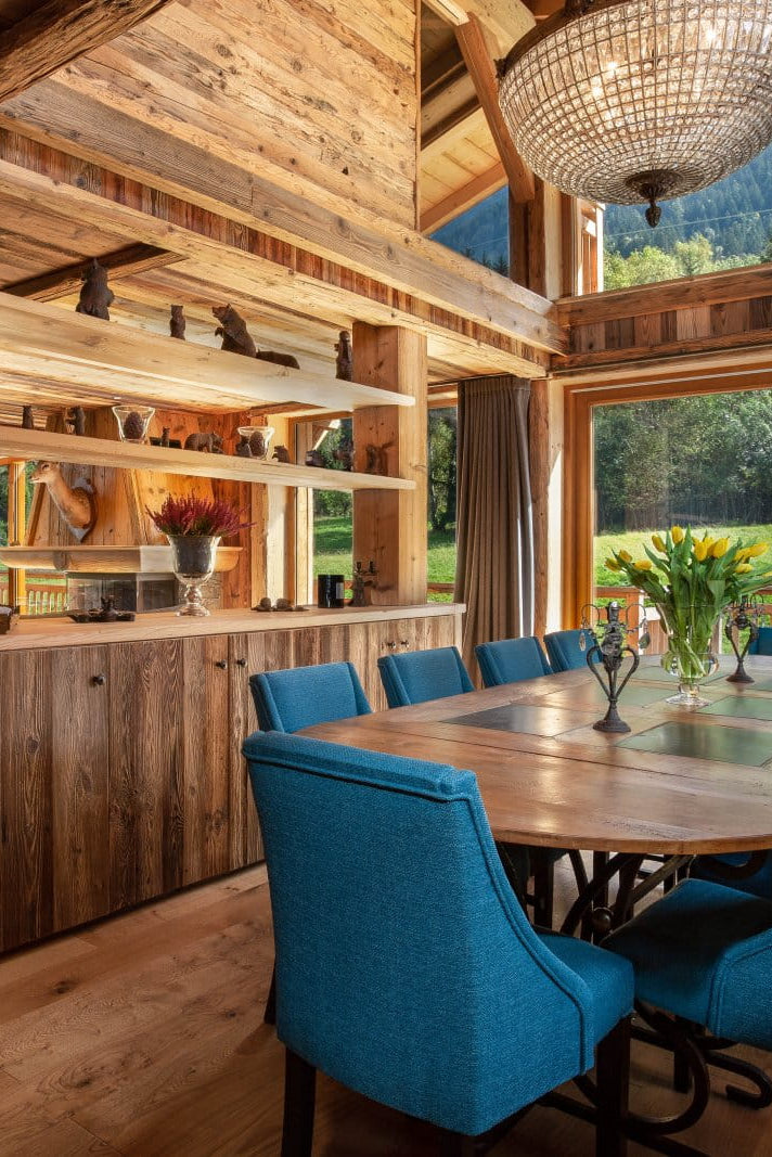 A rustic wooden chalet interior featuring a dining area with a rectangular wooden table and eight teal-blue upholstered chairs, a wooden bar counter with open shelving, and large windows overlooking a green mountain landscape.