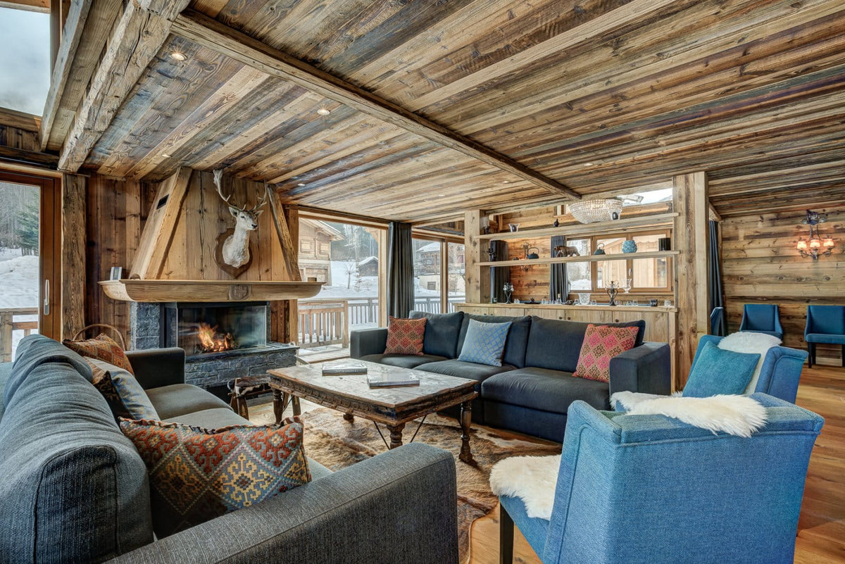 A rustic mountain chalet living room with a large stone fireplace, wood-paneled ceiling, blue sofas, a wooden coffee table, and a blue armchair with a white faux-fur throw.
