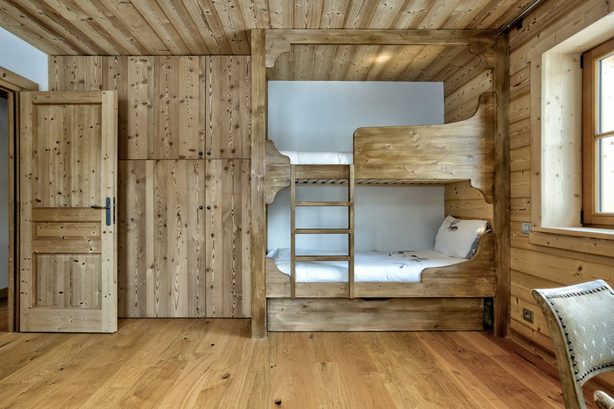 A rustic wooden bunk bed with a built-in ladder, set against a wood-paneled wall in a cabin-style bedroom with matching wood floors and door.