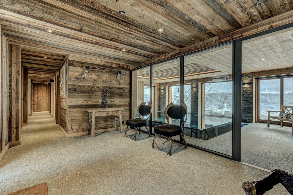 A rustic interior space with exposed wood ceiling and wall paneling, beige carpet, two black chairs near large floor-to-ceiling glass windows, and a hallway on the left.