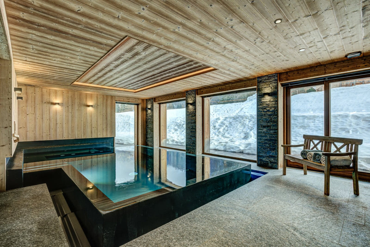 A luxury indoor ski resort spa featuring a modern rectangular plunge pool with clear blue water, set in a rustic wooden chalet with large windows overlooking a snowy mountain landscape.