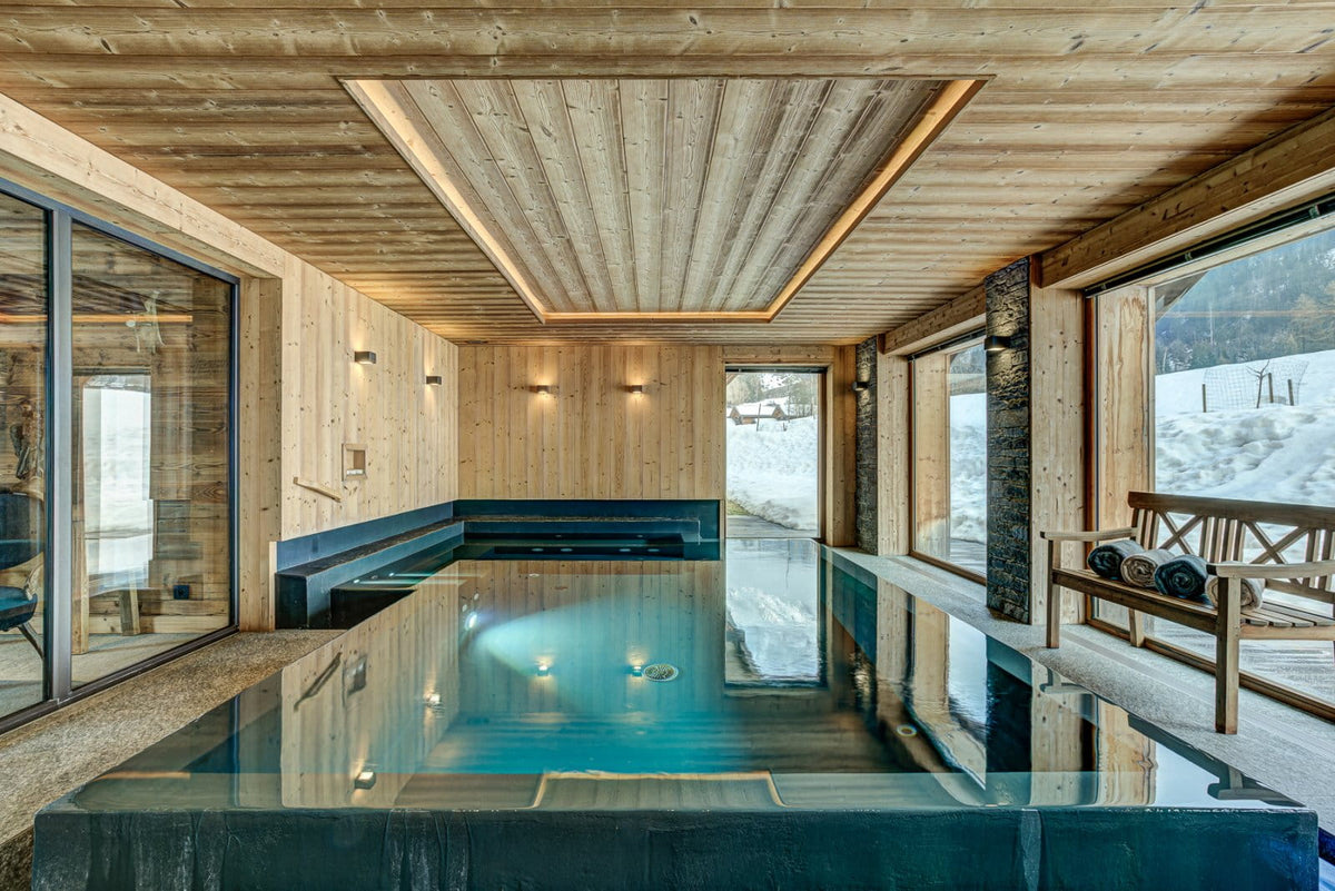 A luxury indoor mountain pool with clear blue water, surrounded by light wood walls and ceiling, with a wooden bench and large windows showing a snowy outdoor landscape.