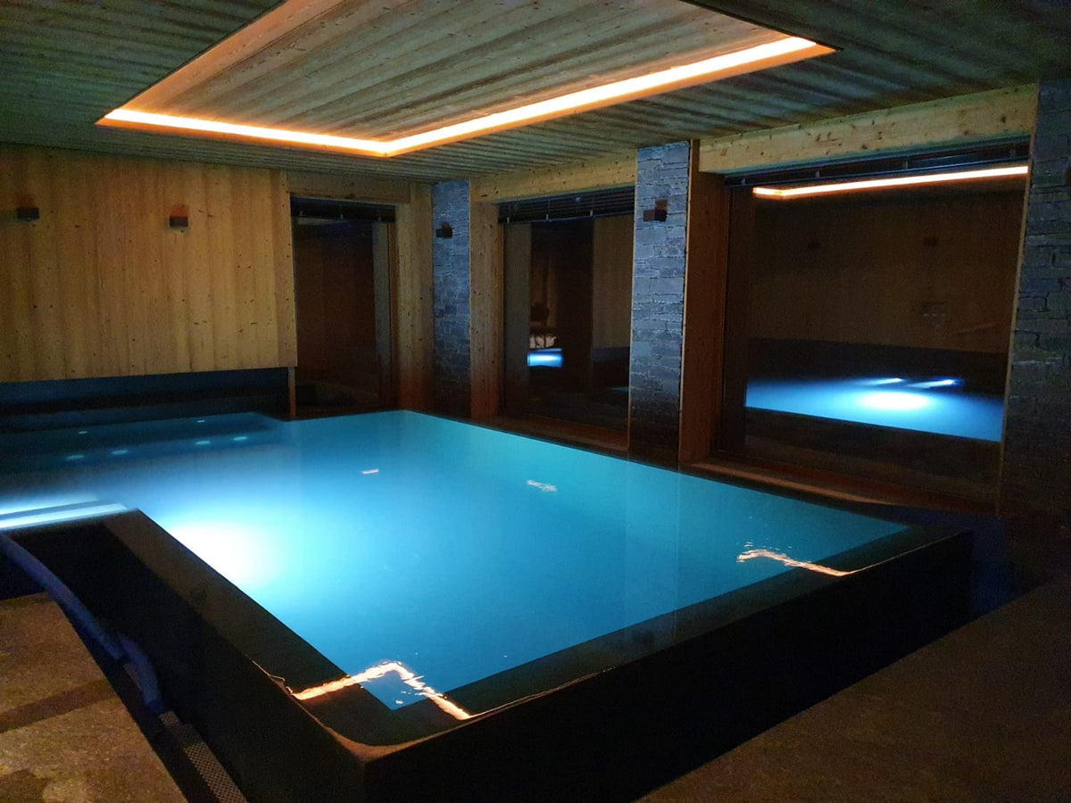 An indoor heated pool or spa basin with glowing blue water, surrounded by wood-paneled walls and ceiling with recessed lighting.