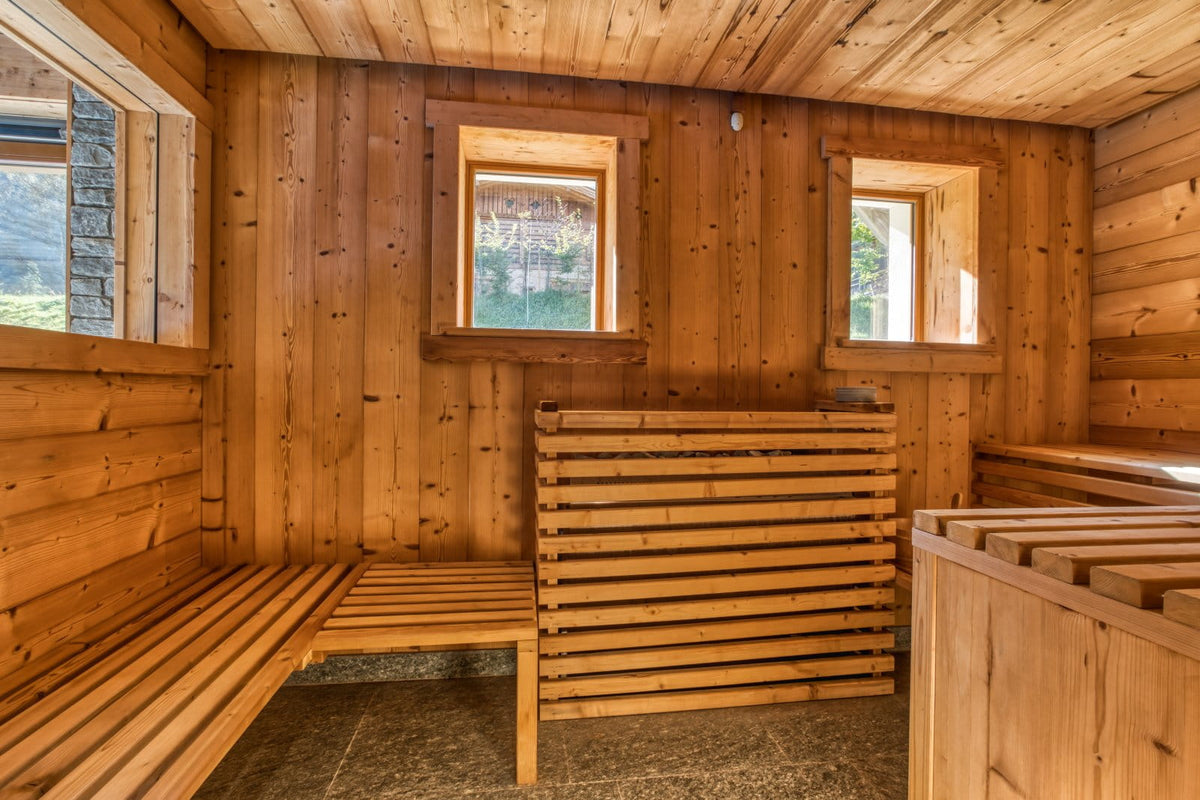 A rustic wooden sauna interior with slatted benches, paneled walls and ceiling, and small windows showing outdoor scenery.