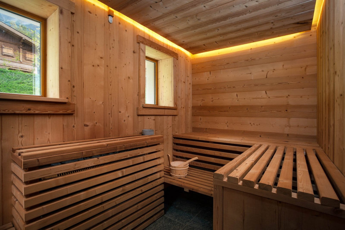 A traditional wooden sauna interior with tiered wooden benches, wood-paneled walls and ceiling, warm yellow LED strip lighting, and a small hanging bucket with a wooden scoop near one bench.