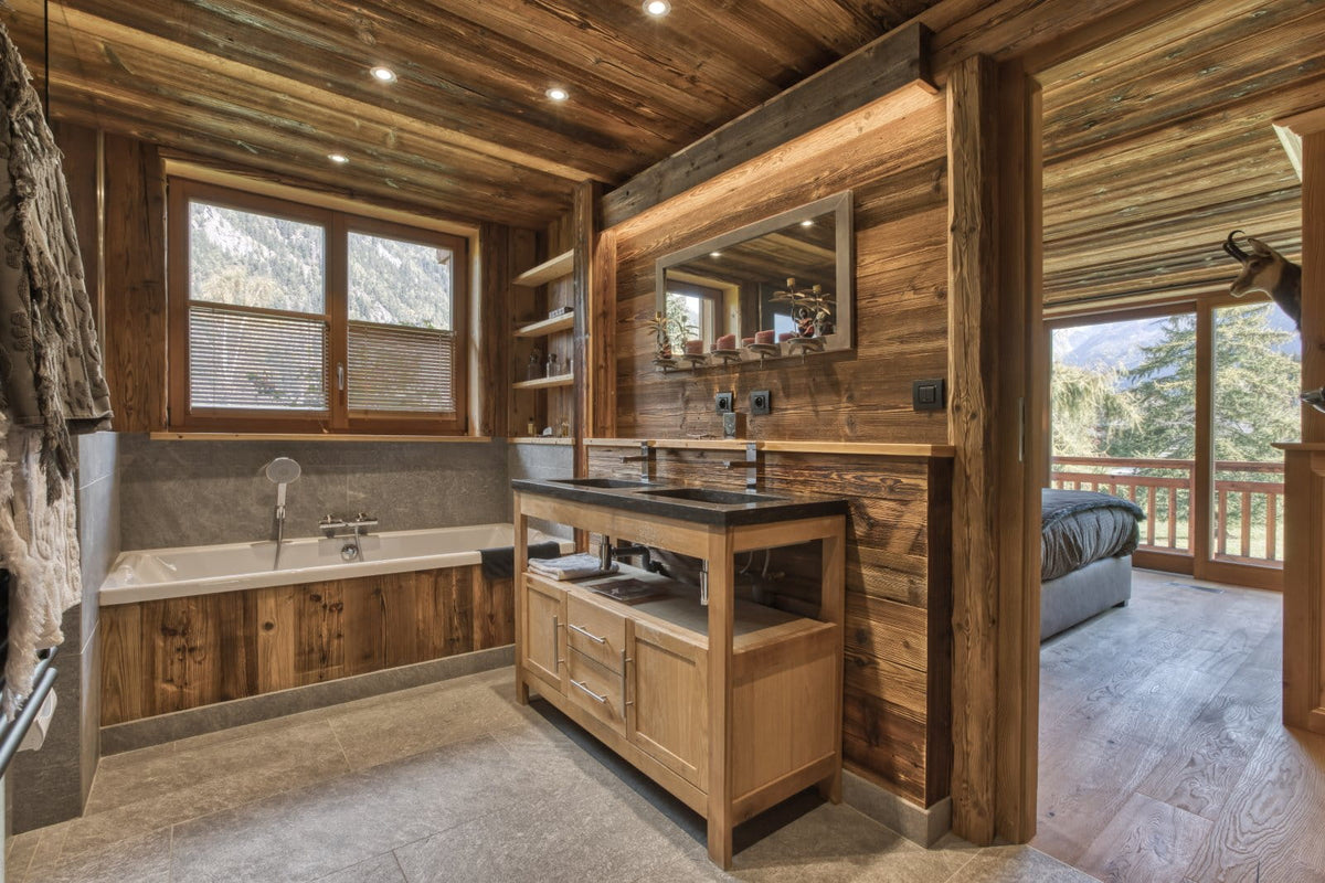 A luxury rustic bathroom interior with a wooden vanity, double sinks, a stone bathtub, and large windows overlooking a mountain landscape.