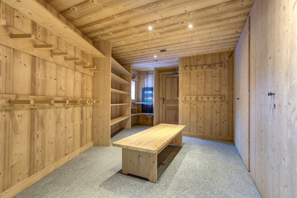 A spacious wooden storage room with light natural wood walls, ceiling shelves, a matching wooden bench, and gray carpeted flooring.