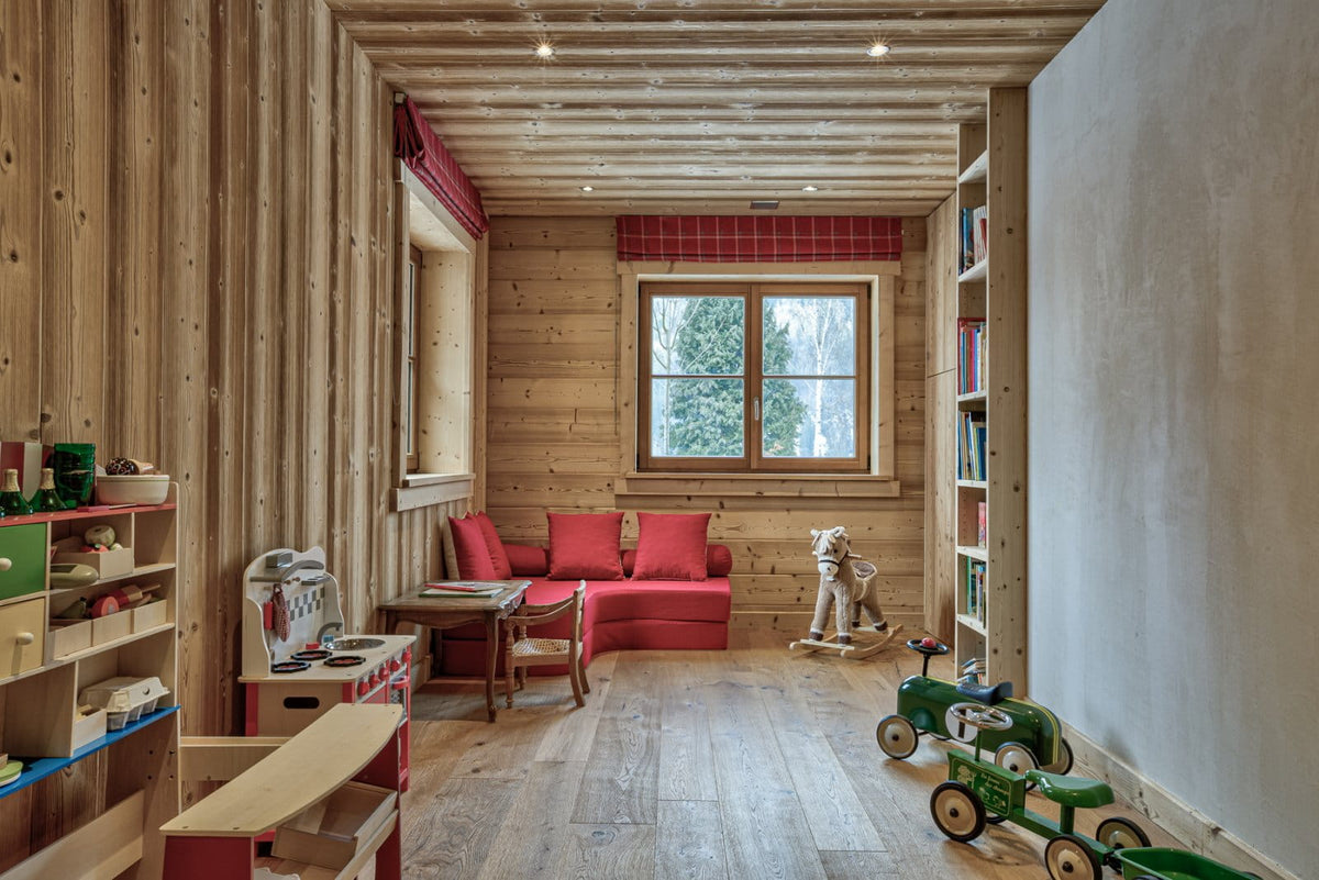 A cozy children's playroom with light wood paneled walls and ceiling, featuring a red sofa, a rocking horse, green toy cars, and wooden shelves.