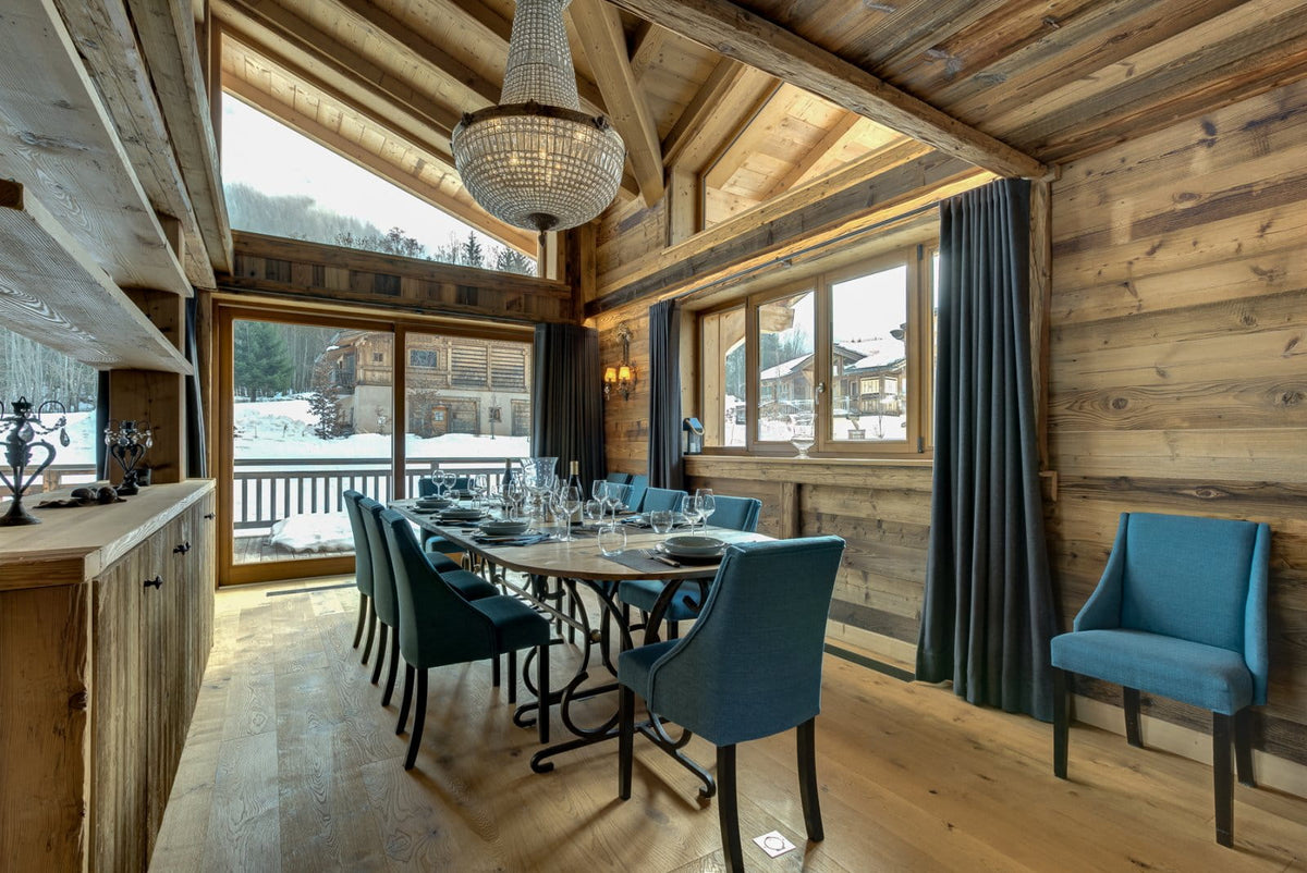 A luxury ski chalet dining room featuring a round wooden dining table set with teal blue upholstered chairs, a large crystal chandelier, and a view of a snowy mountain landscape through the windows.
