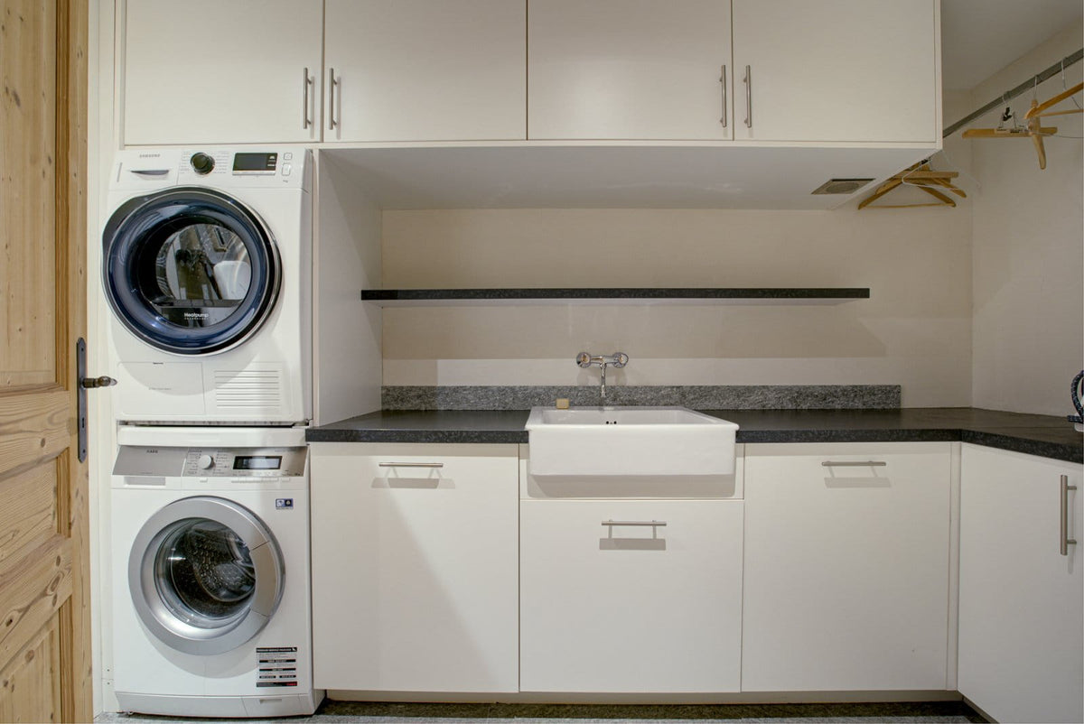 A compact laundry room setup with a stacked white washer and dryer on the left, white cabinets, a white farmhouse sink, dark countertops, and a hanging rod with clothes hangers on the right.