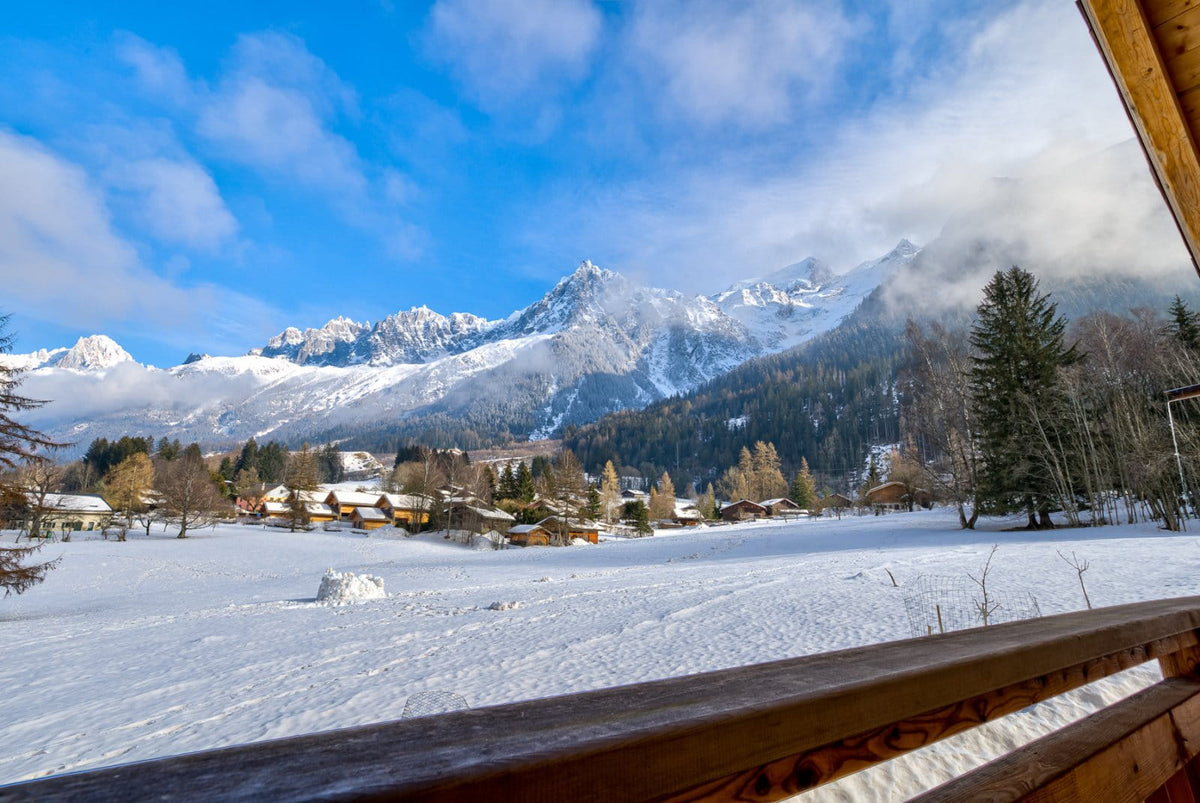 A scenic winter mountain landscape viewed from a wooden balcony, with snow-covered ground, small alpine cabins, evergreen trees, and jagged snow peaks under a bright blue sky with white clouds.
