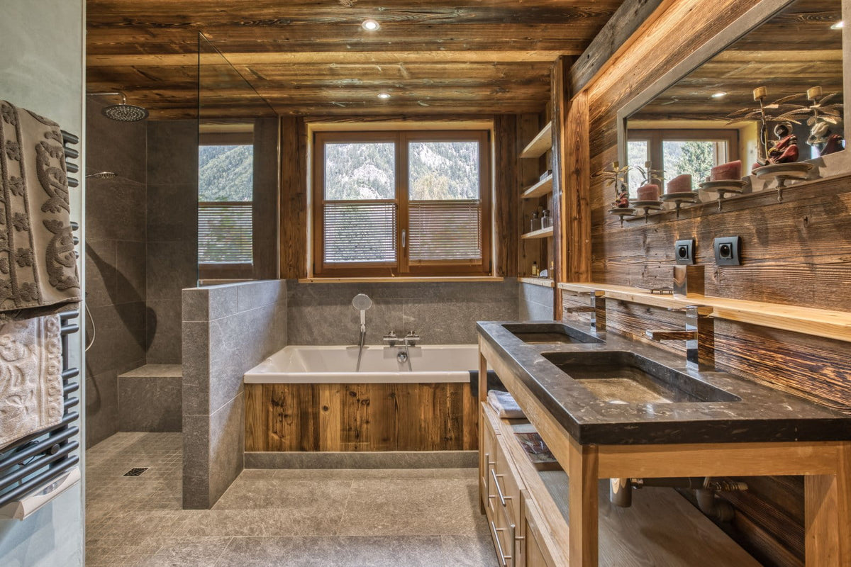 A luxury rustic bathroom interior featuring a double vanity with a dark stone countertop and wood cabinetry, a built-in bathtub with wood paneling, a glass shower enclosure, and a large wood-paneled wall mirror.