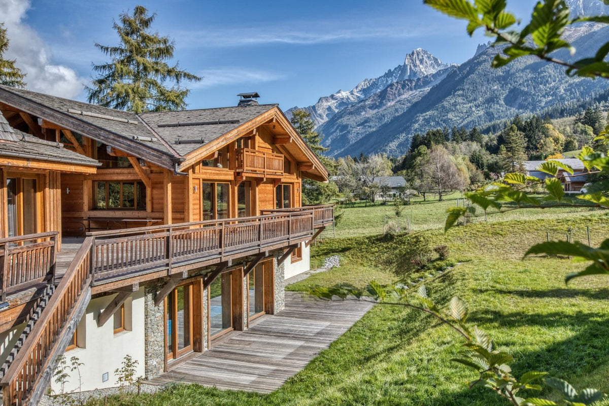 A large alpine-style wooden chalet with balconies and a wooden deck, set in a grassy field with mountains and trees in the background under a blue sky.