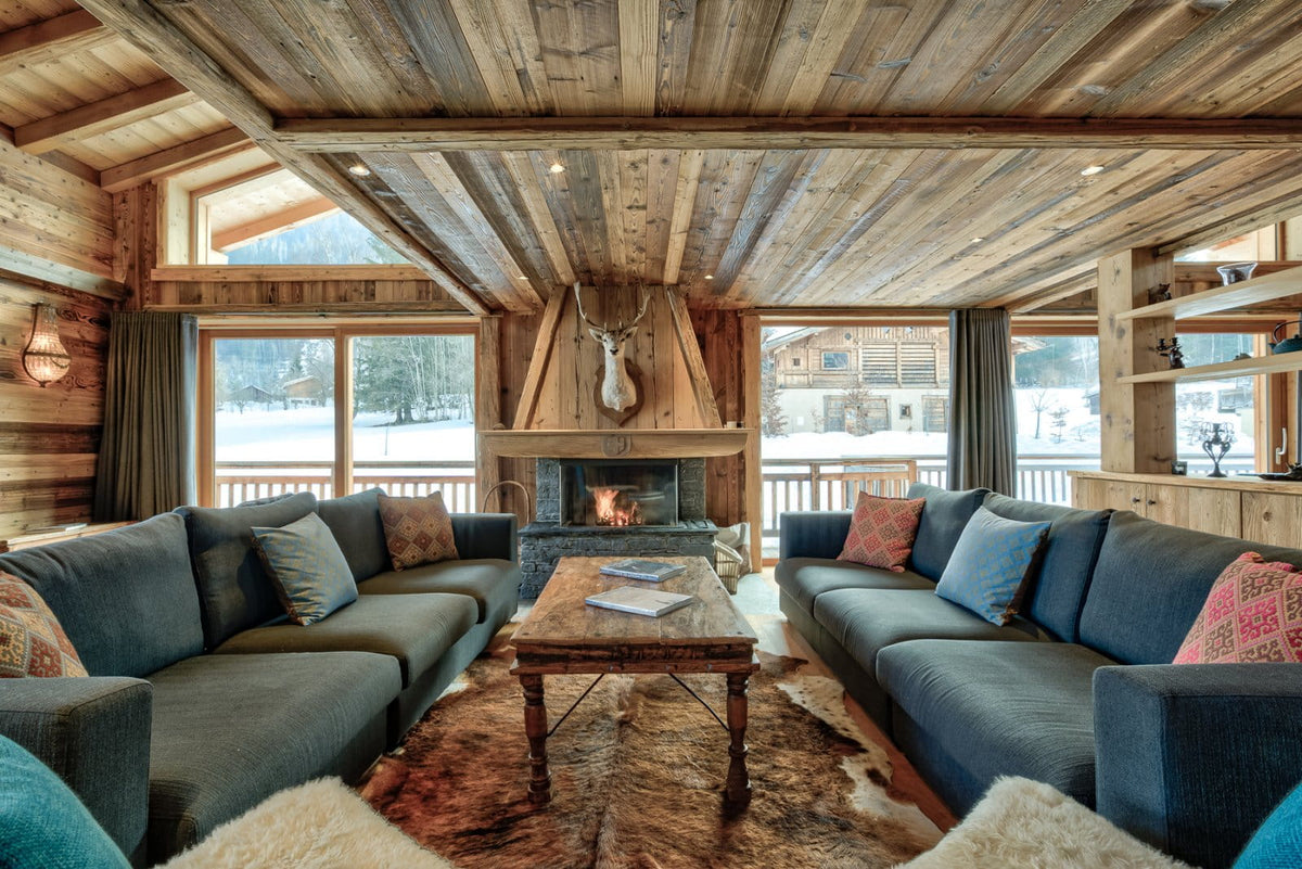 A cozy ski chalet living room with dark blue sectional sofas, a rustic wooden ceiling, a stone fireplace with a fire burning, and large windows looking out onto a snowy outdoor landscape.