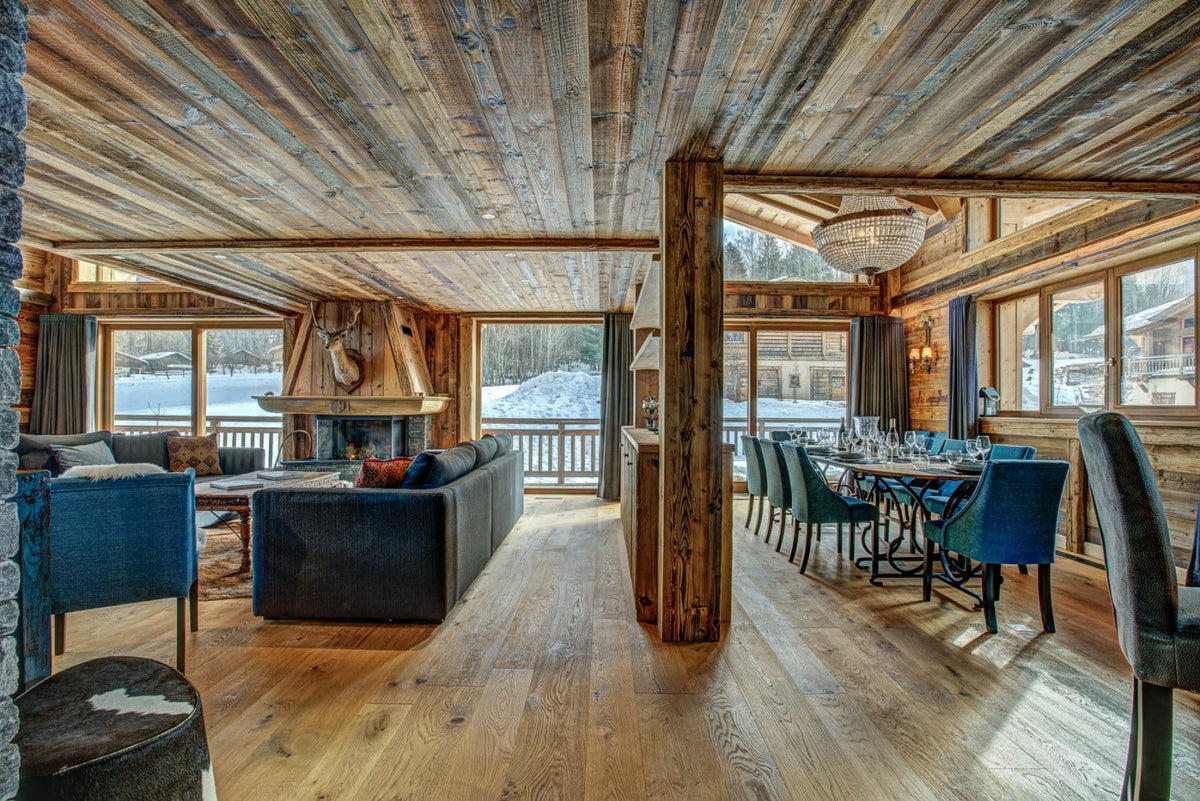 A luxurious ski chalet interior featuring a high-ceilinged rustic wood ceiling, hardwood floors, a dark leather sofa in the living area, and a dining set in the adjacent room, with snowy outdoor views visible through large windows.