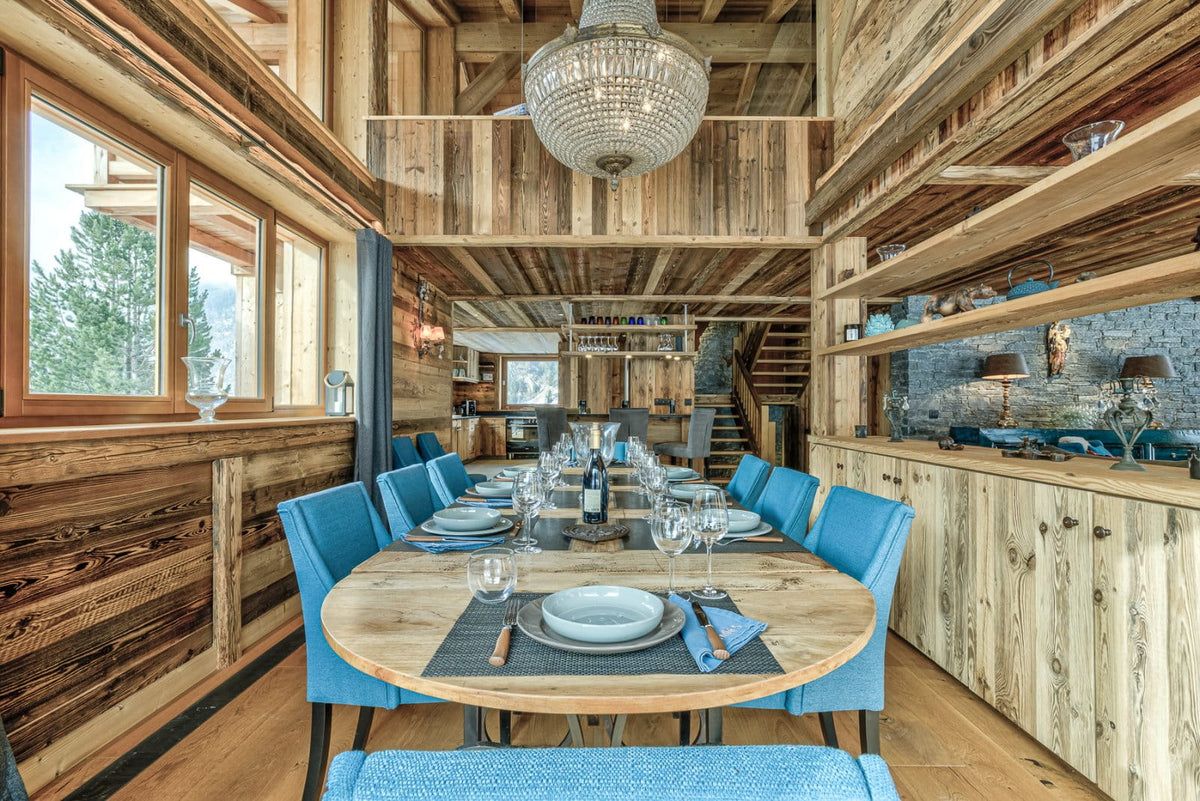 A luxury dining room with a round wooden table set for six with blue upholstered chairs, set against rustic wood-paneled walls and a loft staircase in the background.