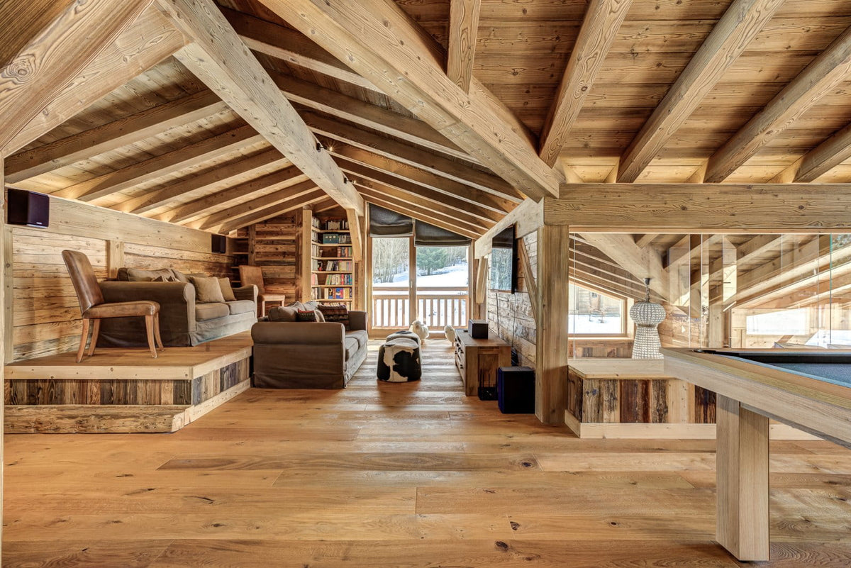 A large rustic wooden chalet interior with exposed timber ceiling beams, hardwood floors, built-in bookshelves, a living room with sofas, and a pool table in the foreground.