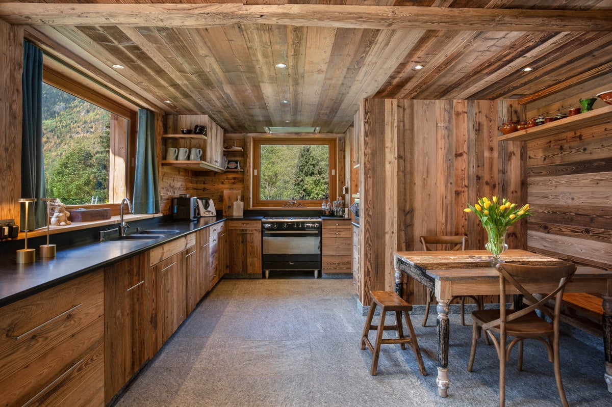 A rustic wooden chalet kitchen and dining area with exposed wood ceiling beams, dark countertops, built-in cabinetry, a stove, and a wooden dining table with chairs holding yellow flowers.