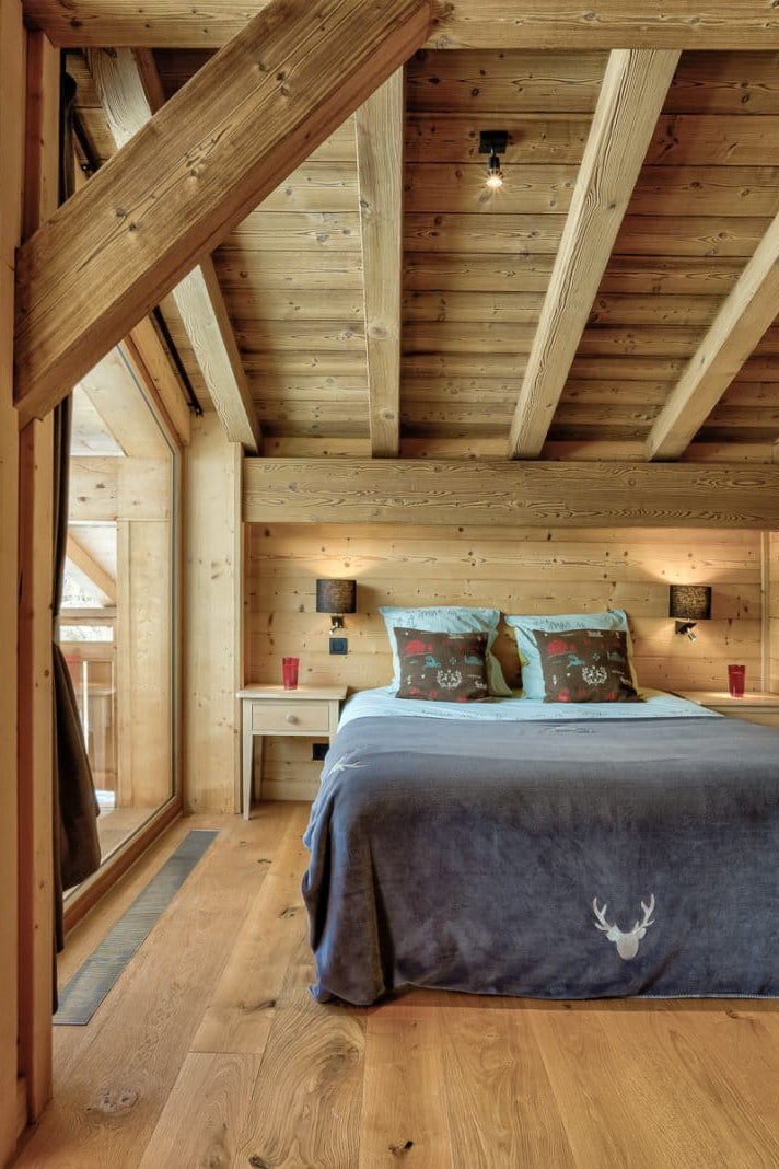 A cozy bedroom interior with a bed dressed in a dark blue blanket and patterned pillows, set in a rustic wooden chalet with exposed beams, wood-paneled walls, and large windows overlooking a mountain view.