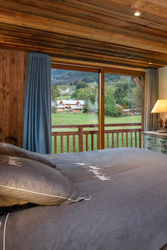 A cozy bedroom interior with a large bed dressed in gray bedding, set in a rustic wooden cabin with a fireplace, blue curtains, and a window overlooking a green outdoor landscape.