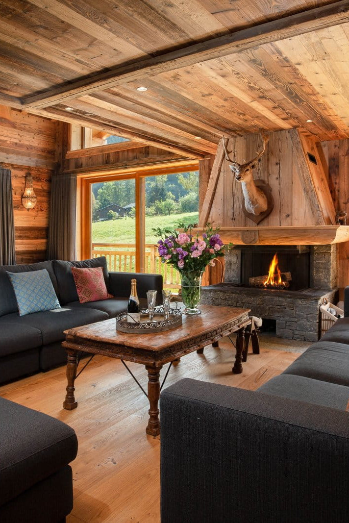 A luxurious mountain chalet living room featuring dark gray sectional sofas, a rustic wooden coffee table, a stone fireplace with a large wood-burning chimney, and large windows looking out onto a green valley.