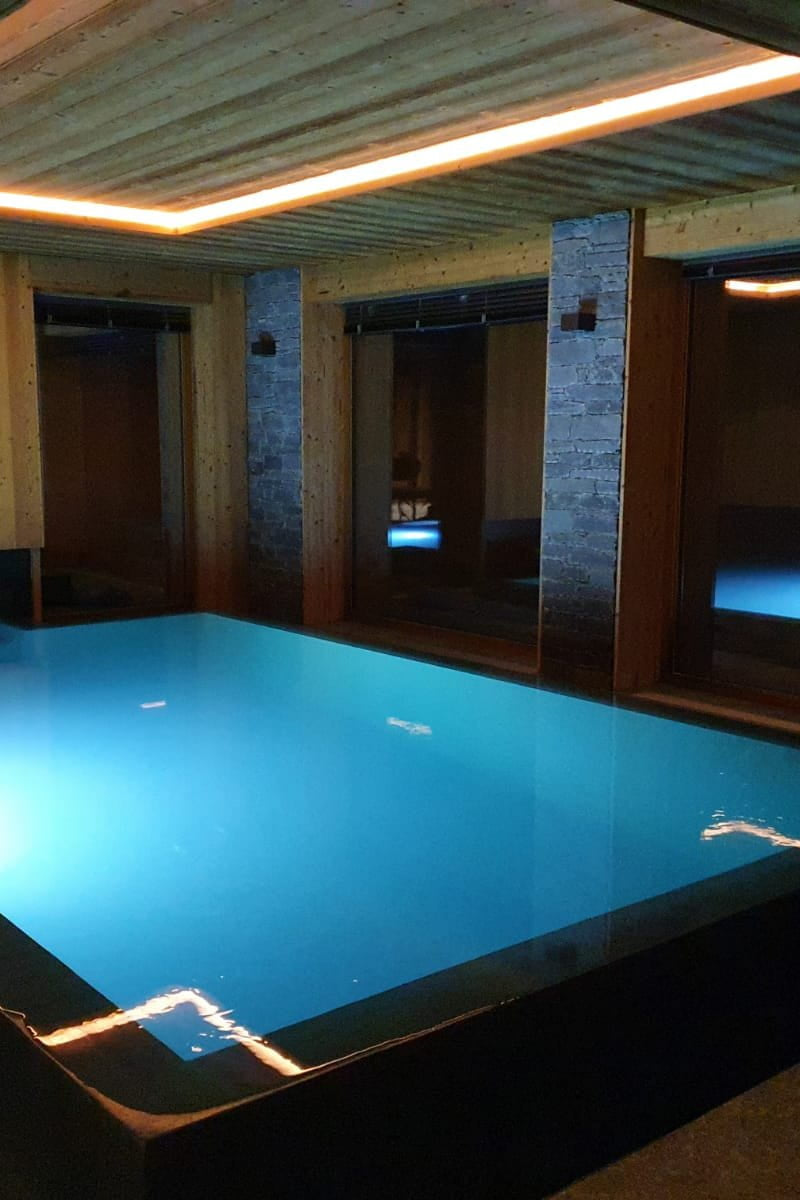 An indoor heated pool or spa basin with glowing blue water, surrounded by wood-paneled walls and ceiling with recessed lighting.