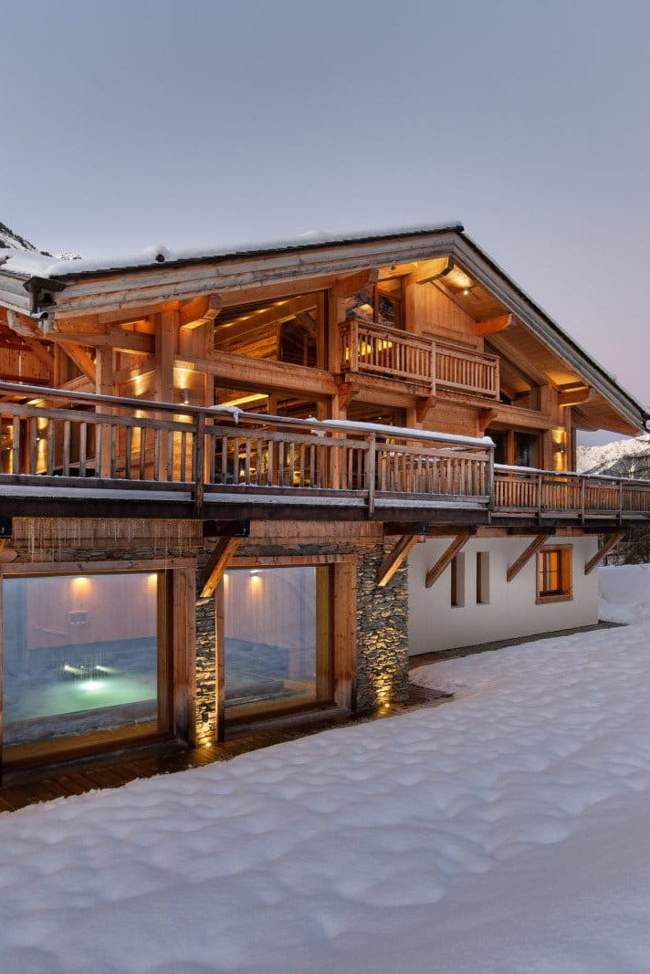 A luxury alpine chalet with warm glowing windows and wooden balconies, nestled in a snowy mountain landscape at dusk.