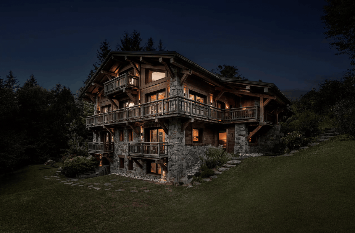 A large alpine-style chalet house at night with warm glowing windows and balconies, set on a grassy hillside with dark trees around it.