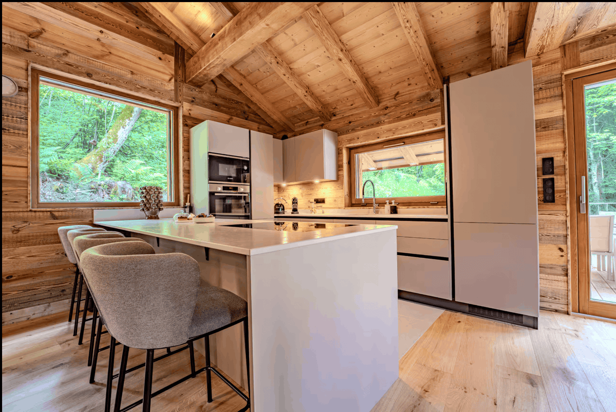 A modern kitchen island with a white countertop and base, surrounded by four upholstered bar stools with black metal legs, set in a rustic wood cabin with a sloped wooden ceiling.