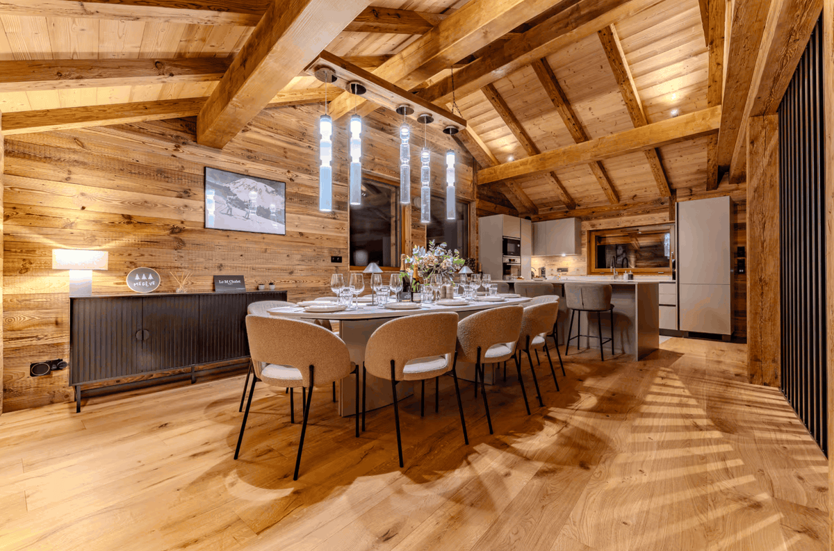 A modern luxury chalet interior featuring a large dining area with a round table, six beige upholstered chairs, and a long dining table set with glassware. The room has a high wood-beamed ceiling, warm wood-paneled walls, and a contemporary kitchen in the background.