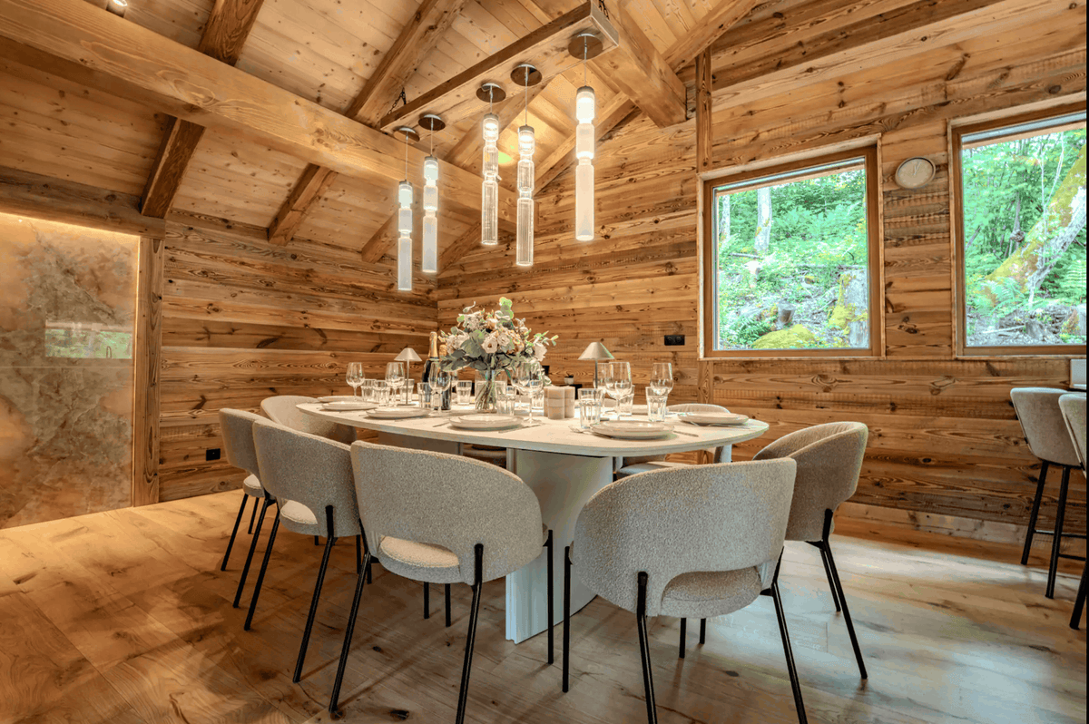 A modern dining room with a round white table set for six, surrounded by six light gray upholstered chairs with black legs. The room features warm wood-paneled walls and ceiling, with a multi-light pendant fixture hanging above the table.