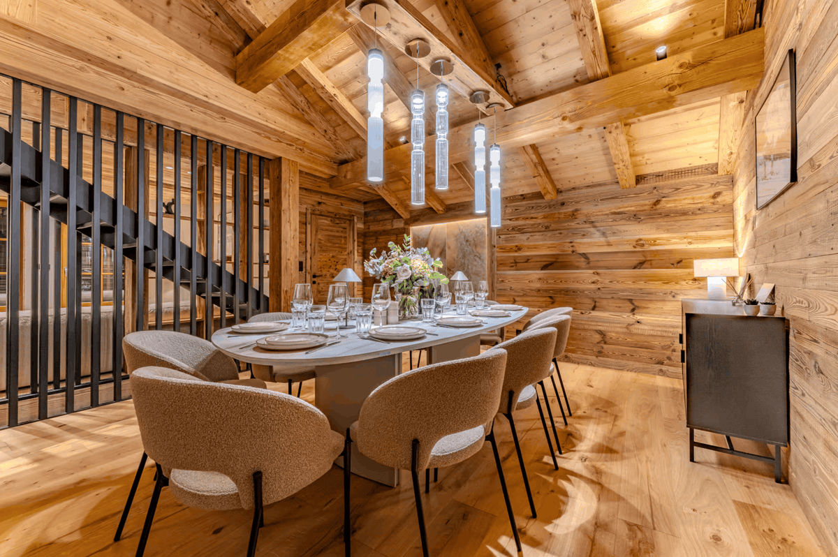A luxury dining room with warm wood-paneled walls and ceiling, featuring a long oval table set with glassware and a floral centerpiece, surrounded by modern upholstered dining chairs. A black metal staircase railing is visible on the left, and cylindrical pendant lights hang from the ceiling.
