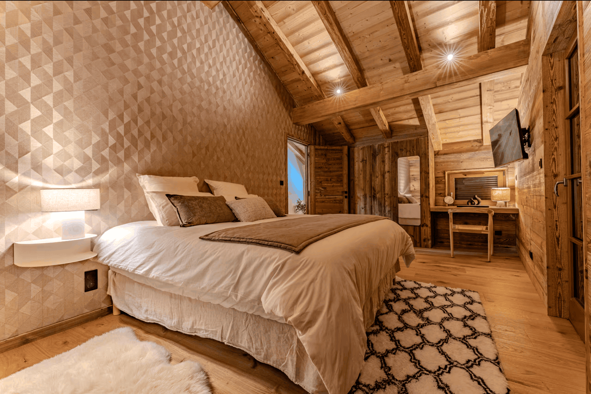 A cozy, rustic bedroom with a large bed dressed in cream bedding, set in a wooden chalet-style room with geometric patterned wallpaper, exposed wood ceiling beams, and a desk area in the background.