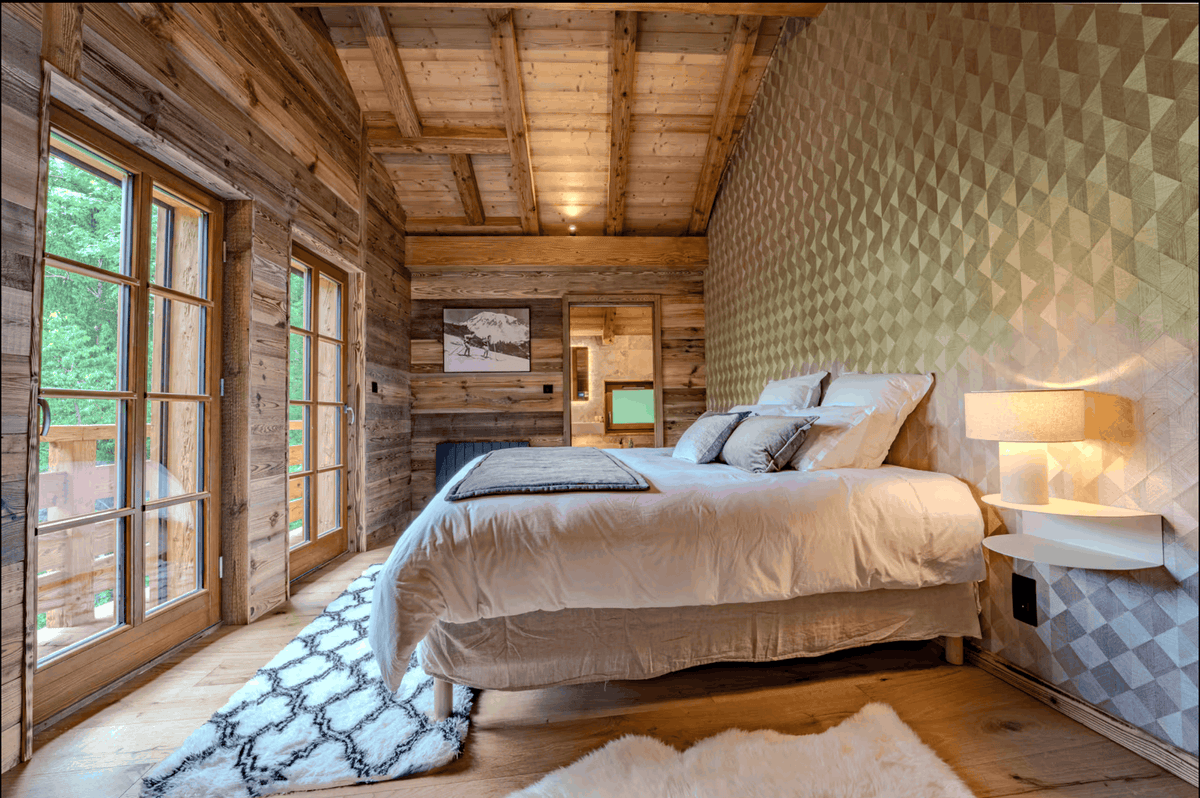 A rustic mountain chalet bedroom featuring a large bed with white bedding, a geometric patterned accent wall, wood-paneled ceiling, and large windows looking out to green trees.