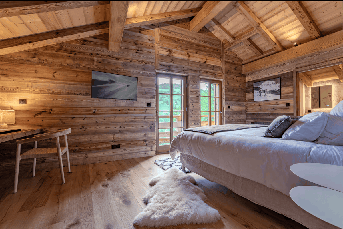 A rustic mountain chalet bedroom featuring a large bed with white bedding, a wooden desk and chair, a fluffy white rug, and wood-paneled walls under a sloped wooden ceiling.