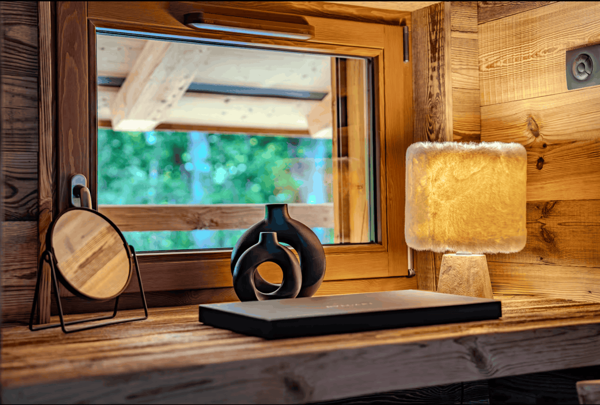 A warm, rustic interior scene with a wooden window, wood-paneled walls, a glowing table lamp with a textured cream shade, a black rectangular object on a wooden surface, and two dark decorative vases. No person or animal is present, but this is not a clean isolated product shot; it is styled in a real environment, so subject_type is Lifestyle.