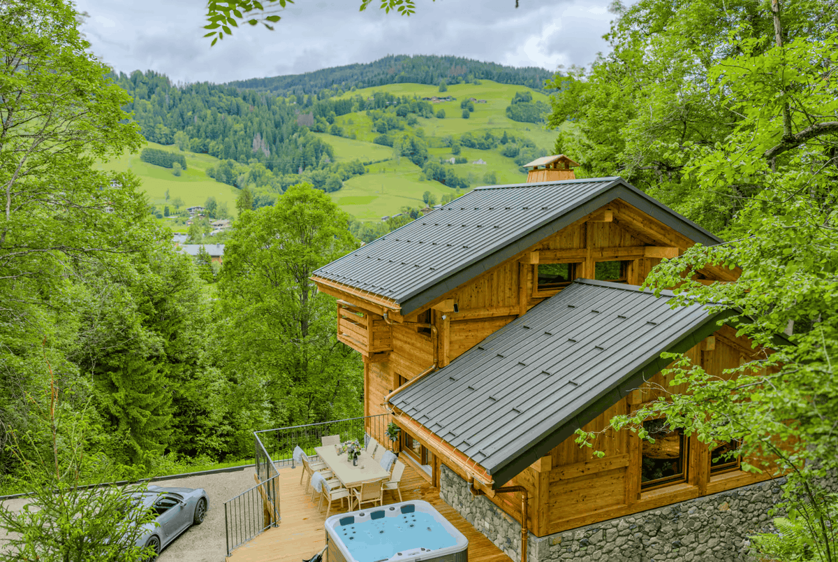 A modern wooden chalet with dark gray metal roofs, situated in a lush green mountain forest. The property features a wooden deck with outdoor dining furniture and a hot tub, with a scenic valley and green hills visible in the background under a cloudy sky.