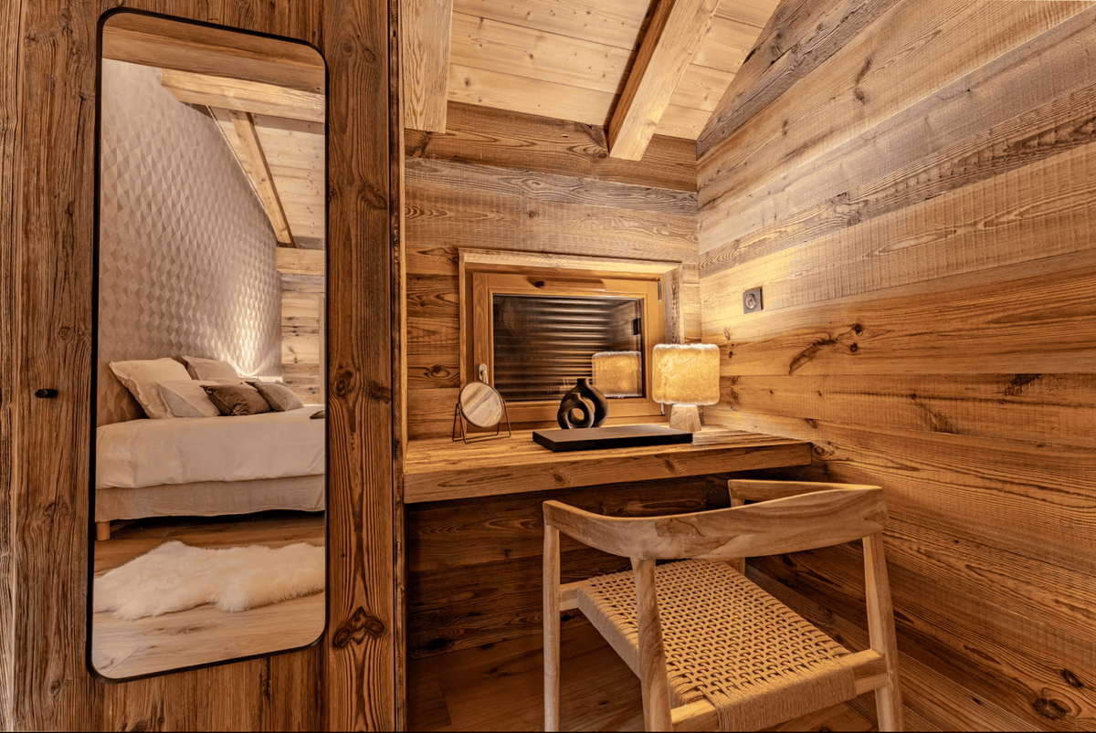 A rustic wooden cabin interior with a built-in desk and mirror, warm wood-paneled walls, a woven chair, and a reflection in the mirror showing a bed area.