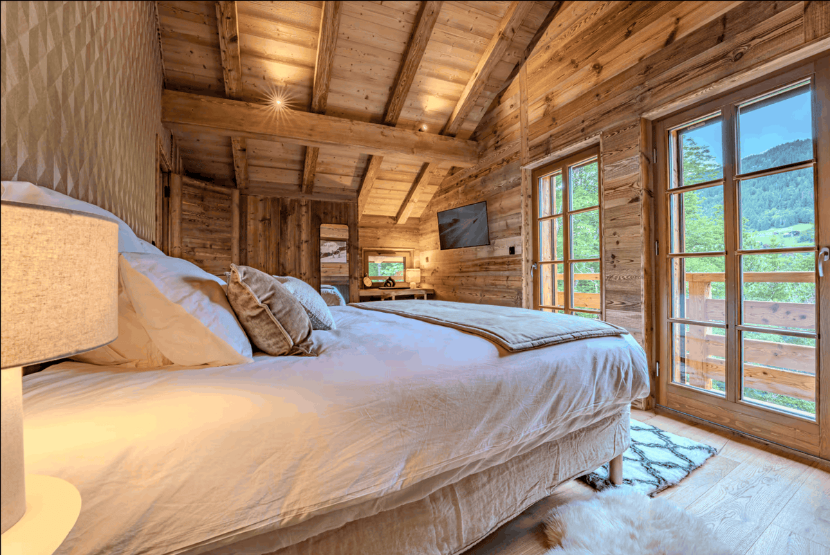 A large bed dressed in white linens and patterned pillows, set in a rustic wooden chalet with wood-paneled walls, exposed wooden ceiling beams, and large windows overlooking a green mountain landscape.