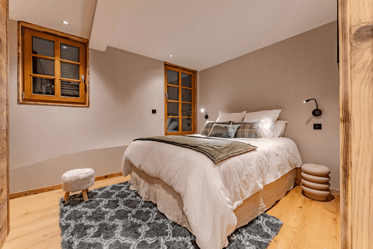 A modern bedroom interior featuring a neatly made bed with white bedding and a gray patterned rug, set against warm beige walls with wooden-framed windows and wall-mounted reading lights.