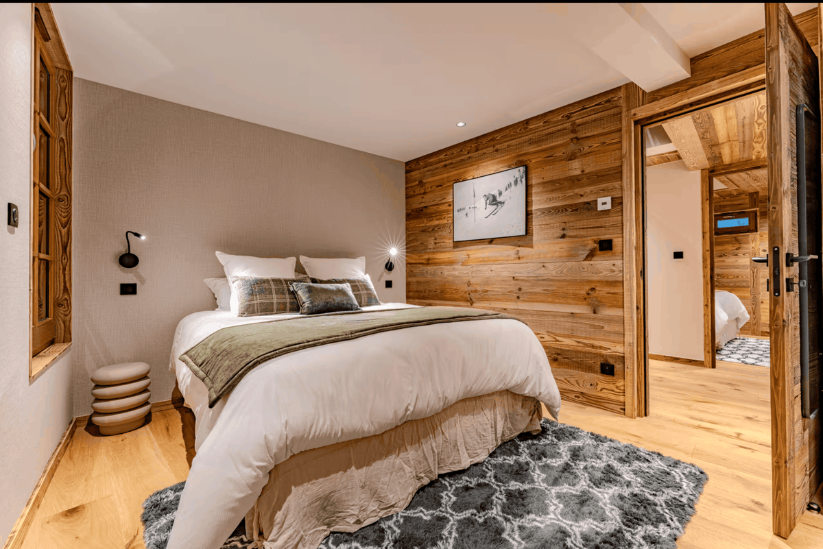 A modern bedroom interior with a neatly made bed dressed in white bedding and a green throw, set against a wood-paneled wall and light wood flooring. A gray patterned rug lies beneath the bed, and an open doorway reveals an adjacent bathroom.