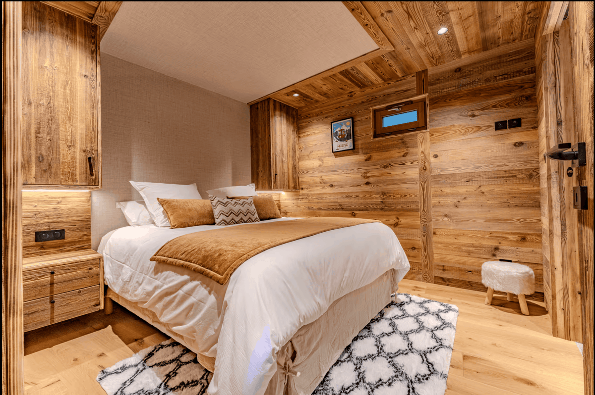 A rustic bedroom interior with a neatly made bed dressed in white bedding and a tan throw blanket, set against wood-paneled walls and ceiling. A patterned rug lies at the foot of the bed, with a small stool and framed wall art visible in the room.