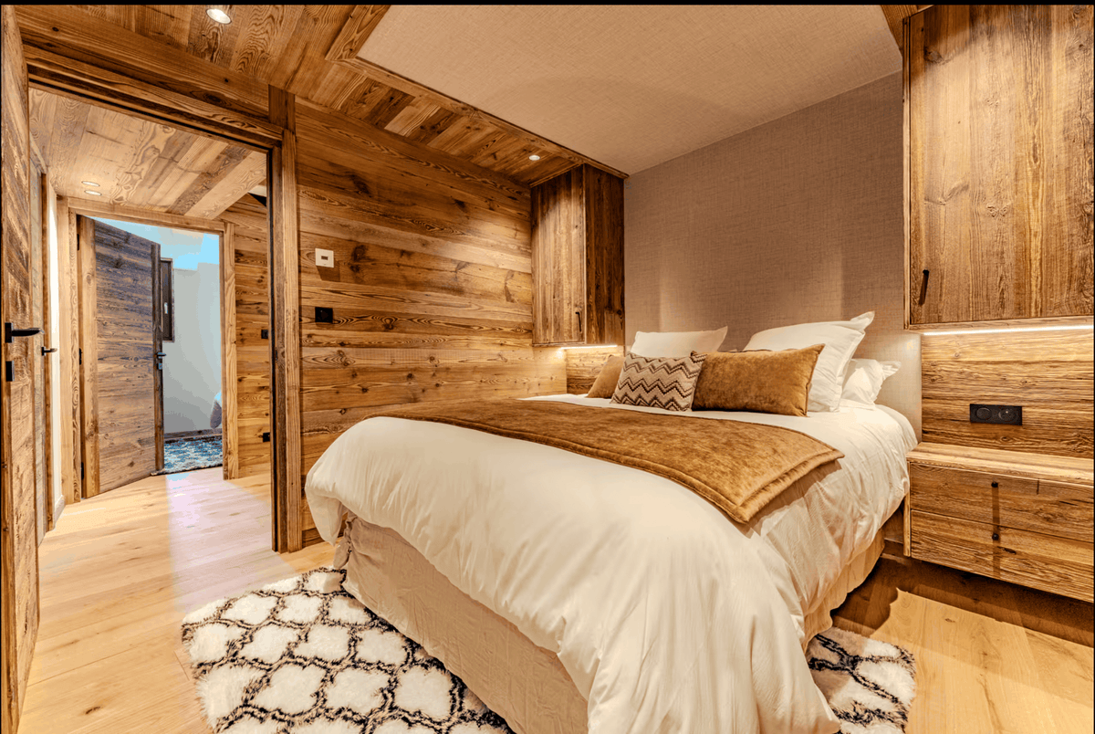 A cozy bedroom interior with a neatly made bed dressed in white bedding and a tan throw blanket, set against rustic wood-paneled walls and matching wood furniture. A patterned rug is visible at the foot of the bed, and an open doorway on the left reveals another room.