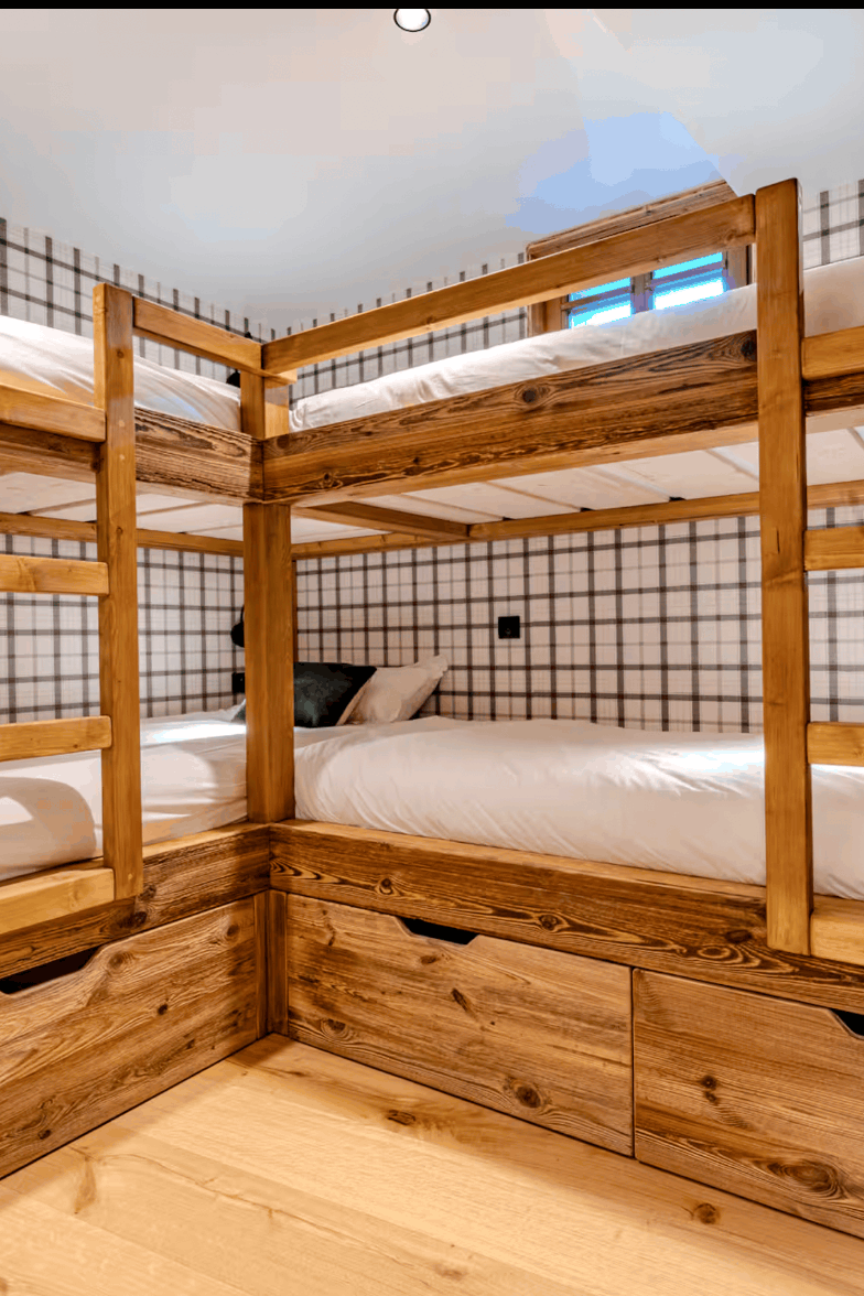 A rustic wooden bunk bed frame with three single beds, white bedding, and built-in storage drawers underneath, set against a black and white checkered wall.