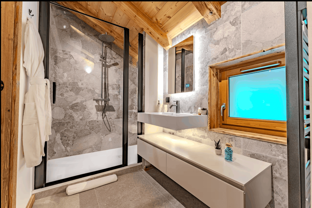 Modern luxury bathroom featuring a large walk-in shower with a glass door, a floating white vanity with a sink, a wood-framed window displaying a bright blue screen, and a white robe hanging on the left.