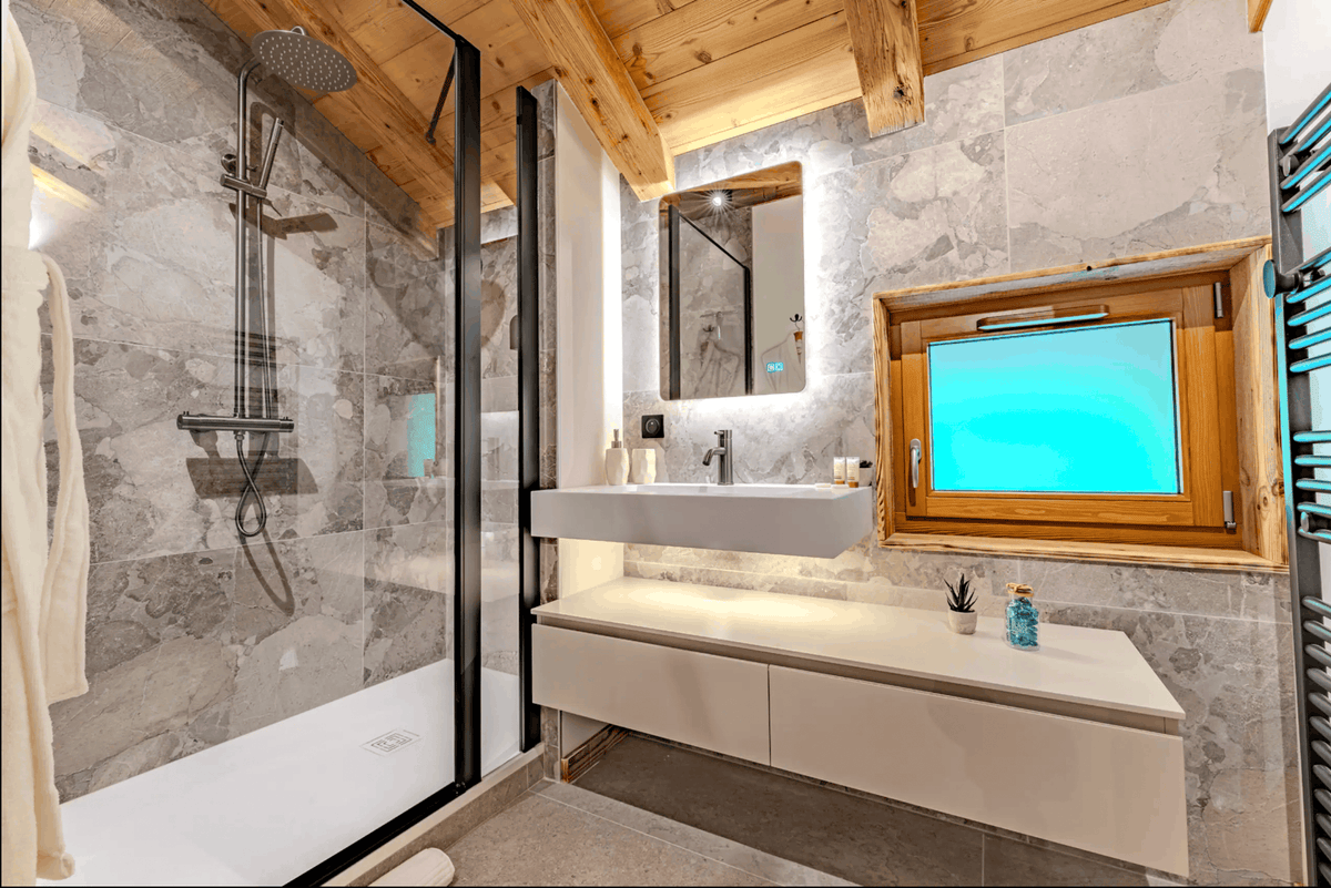 Modern luxury bathroom with a large walk-in shower, floating white vanity, gray stone tile walls, exposed wood ceiling beams, and a built-in wood-framed TV screen.